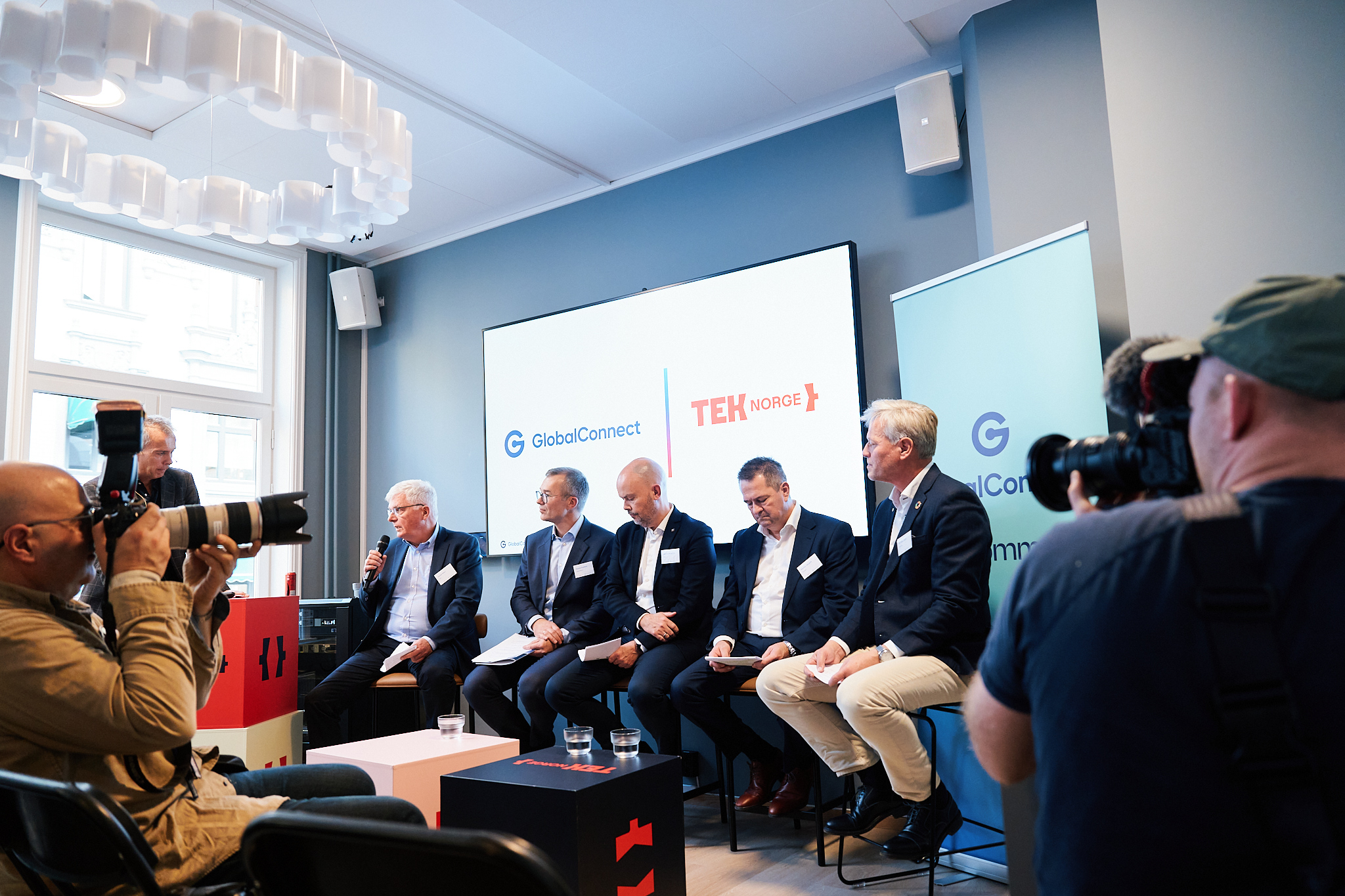 Paneldeltakere under fremleggelsen av rapporten til Global Connect. Sittende på barkrakker foran presentasjonsskjerm. Presse til stede.